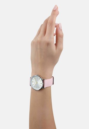 Runde Uhr mit silbernem Ziffernblatt, goldenen Markierungen und pinkem Silikonarmband. Die Zeiger zeigen die Zeit an, das Handgelenk ist vor einem weißen Hintergrund sichtbar.