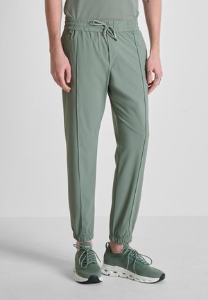 Hombre con pantalones jogger verde claro con cordón y zapatillas a juego verde claro con suelas blancas, de pie sobre un suelo gris claro.