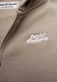 Beige zip-up hoodie gjord av mjukt tyg med ribbad textur; har vitt broderat märkesnamn "Band of Rascals" på bröstet.