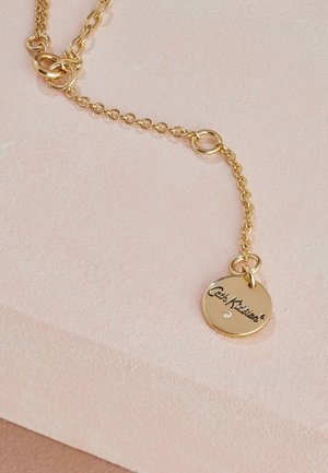 Gold chain necklace with round pendant engraved "Cath Kidston" and a small embedded gemstone, displayed on a soft pink textured surface.