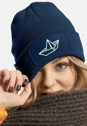 Navy blue knitted beanie with a textured finish and a white paper boat graphic on a rectangular patch.
