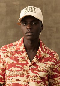 Casquette beige avec logo "LES DEUX" et une chemise à manches courtes à motifs de paysage dans des tons jaune et beige.