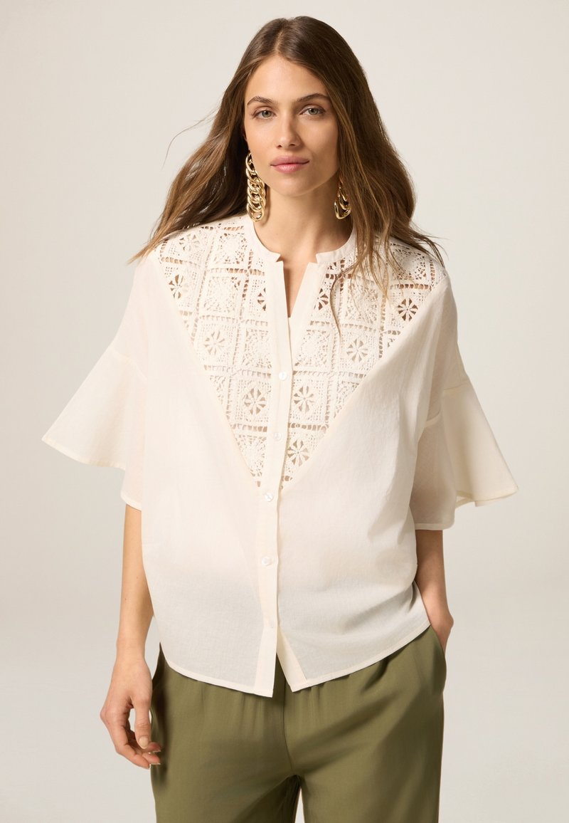 Blusa crema con dettagli in pizzo sulla parte anteriore, con chiusura a bottone e maniche corte a campana. Modello abbinato a pantaloni verde oliva.