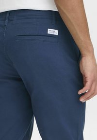 Pantalones azul marino fabricados en mezcla de algodón. Presentan un bolsillo trasero con etiqueta. Textura suave, corte recto y elementos de diseño mínimos.