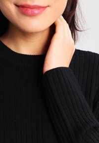 Mustakangas neulepaita, jossa on pyöreä pääntie, tiukat hihansuut ja teksturoitu kangas. Iho näkyy lähellä pääntietä ja käden kohdalla.