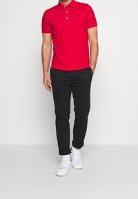 Czerwona koszulka polo z kołnierzykiem i trzema guzikami, w zestawieniu z czarnymi spodniami o slim fit i białymi trampkami. Gładki materiał, casualowy styl.