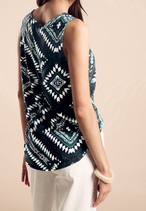 Femme portant un haut noir sans manches avec des motifs géométriques blancs et verts, un pantalon blanc et un bracelet blanc à son poignet.