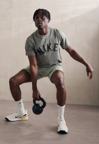 Nike sporta apģērbs, ko valkā cilvēks: olīvzaļš krekls ar melnu logo, gaiši zaļas šortus, baltas zeķes un melna kettlebell.