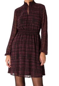 Femme portant une robe à manches longues, de couleur rouge foncé et noir avec un motif, taille froncée et col montant avec une petite ouverture en forme de clé à l'avant.