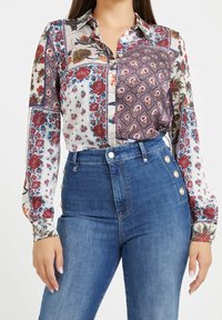 Kvinna i en långärmad blus med lapptäcktsblommönster nedstoppad i högmidjade blå jeans med dekorativa sidoknappar.