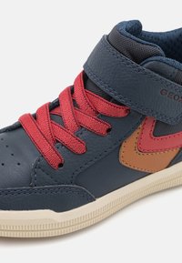 Baskets montantes en cuir bleu marine avec des lacets rouges, présentant un accent marron et une semelle en caoutchouc texturée. Col rembourré pour le confort.