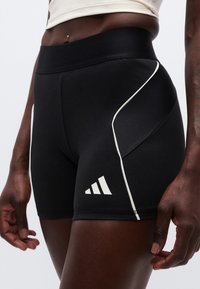 Κοντινό πλάνο ατόμου που φοράει μαύρο αθλητικό σορτς με λευκή επένδυση και λογότυπο Adidas στο αριστερό μηρό, τα χέρια χαλαρά στ ο πλάι.