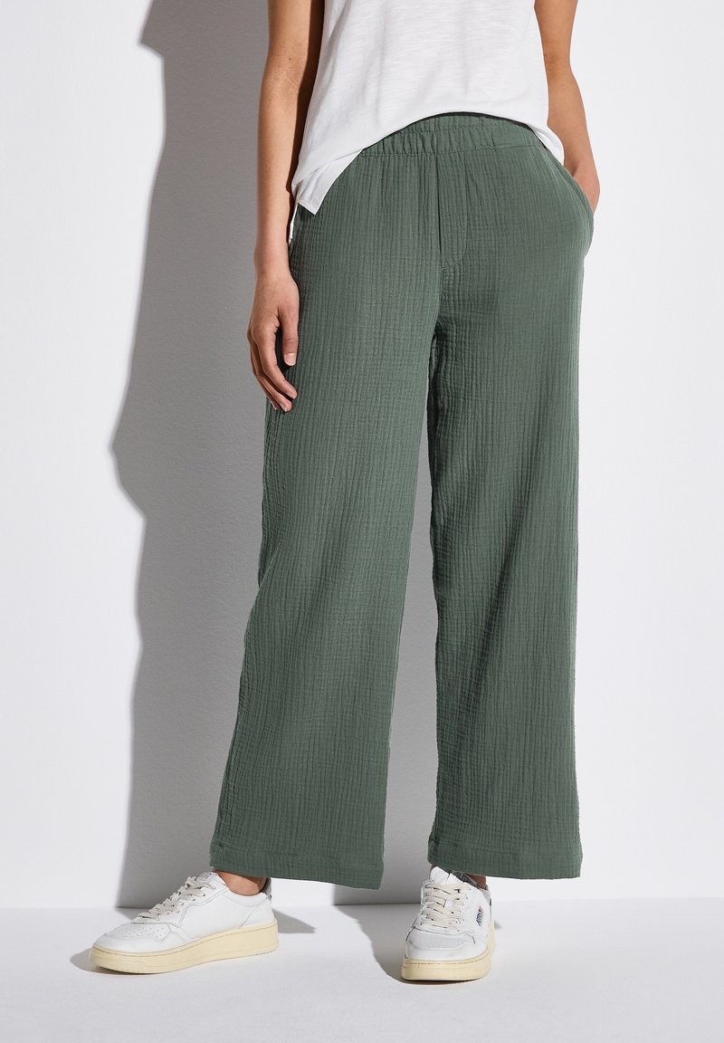 Pantaloni in tessuto verde stropicciato, morbidi e ampi, con elastico in vita e tasche laterali, abbinati a sneakers bianche.