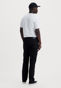 Zwarte denim jeans met een logo op de achterzak, gedragen met een wit t-shirt en een zwarte pet. Het model staat in een neutrale houding.