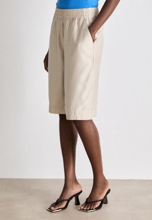 Strak zittende beige kniebroek met fijne strepen, gedragen door een persoon in zwarte sandalen met hak, staand met één hand in de zak.