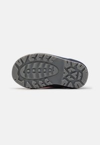 Suela de una zapatilla oscura azul mostrando un patrón de huella detallado con formas ovaladas y geométricas sobre un fondo gris claro.