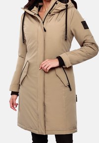 Giacca lunga beige con cappuccio, caratterizzata da dettagli neri, tasche con zip e un tessuto strutturato. Include un logo discreto sul petto.
