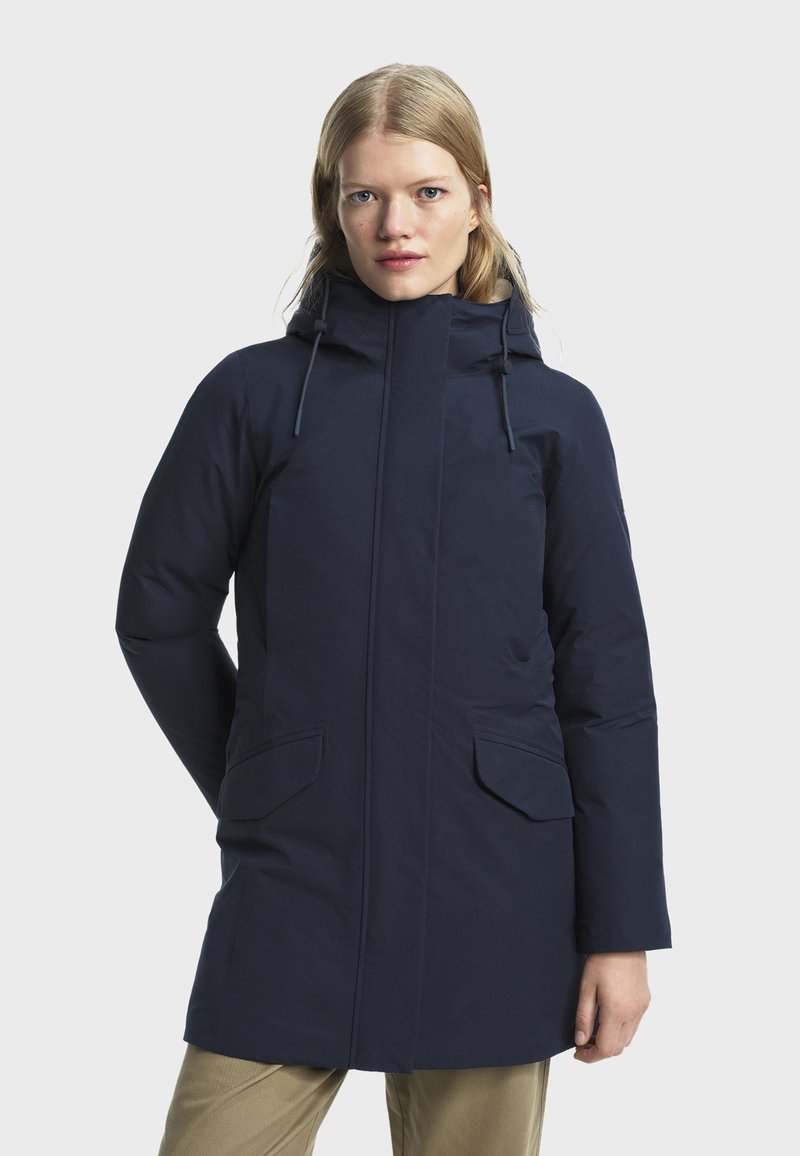 Žena u tamnoplavom zimskom kaputu s kapuljačom i prednjim džepovima, stoji protiv jednostavne svijetle pozadine.