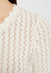 Suéter blanco de punto con un escote en forma de ondas y un patrón texturizado que recuerda a olas o conchas, creando una apariencia ligera.