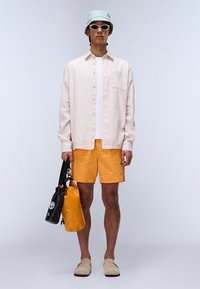 Camisa de botones rosa claro, camiseta blanca debajo, pantalones cortos con estampado naranja, zapatos tipo slip-on beige, gorro de cubo verde menta, sosteniendo bolsas negras y amarillas.