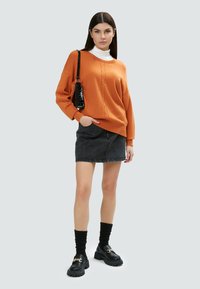 Orange übergroßer Strickpullover, darunter ein weißer Rollkragenpullover, schwarzer Jeansrock, schwarze klobige Loafer und gerippte schwarze Socken.