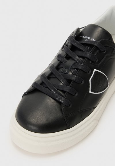 Must musta sneaker, mille on lame paelad, ümar varvas, kontrastne valge tald ja tekstuurne külgaktsent, millel on logo üksikasjad.
