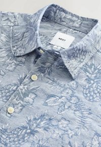 Chemise homme taille moyenne bleu clair avec motifs tropicaux blancs d’ananas et de feuilles et boutons blancs.