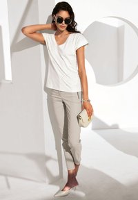 White V-neck t-shirt with embellishments, paired with light grey tapered pants and beige pointed-toe heels, holding a small white clutch.