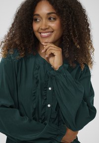 Donna sorridente con capelli ricci, che indossa una blusa verde scuro con volant e bottoni di perla, appoggiando il mento sulla mano e guardando di lato.