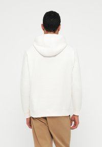 Muž oblečený v obyčajnej bielej mikine s kapucňou a béžových nohaviciach, stojaci chrbtom k fotoaparátu na svetlom pozadí.