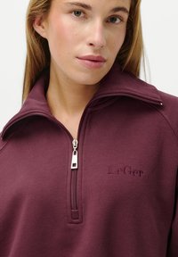 Woman wearing a burgundy zip-up top with a folded collar and "LeGer" embroidered on the chest, close-up view.