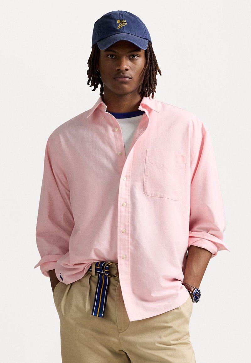 Chemise rose clair à manches longues avec une poche poitrine, portée sur une chemise blanche. Associée à un pantalon beige et une casquette bleue avec un logo jaune.