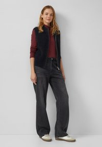 Schwarze Weste mit flauschiger Textur über einem weinroten Langarmshirt, kombiniert mit dunklen weitem Jeans und cremefarbenen Sneakers. Einfaches Design, lässige Passform.