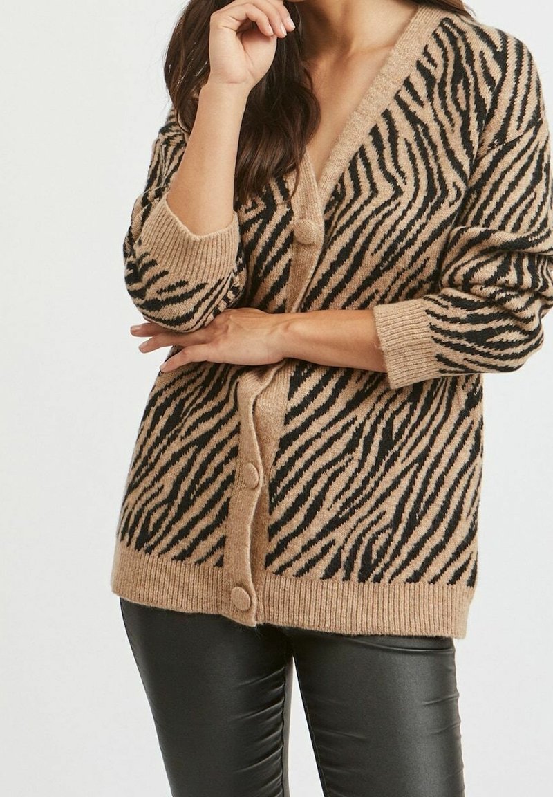 Femme portant un cardigan boutonné beige et noir à rayures zébrées avec des poignets côtelés, associé à un pantalon en cuir noir, posant les bras croisés.