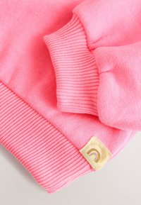 Miękkie różowe mankiety swetra wykonane na drutach z żebrowaną strukturą oraz małą beżową metką z tkaniny z logo w kształcie zakrzywionej linii na jasnym tle.