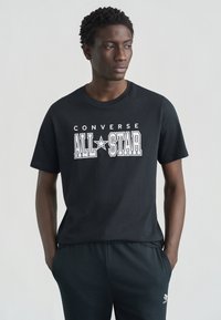 Μαύρο βαμβακερό μπλουζάκι με το γραφικό "CONVERSE ALL STAR" σε λευκό. Μανίκια κοντά, στρογγυλή λαιμόκοψη. Φορεμένο με σκούρα τζόγκερς.