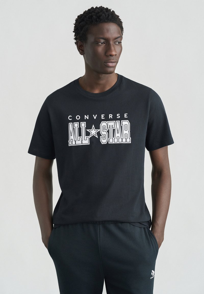 Μαύρο βαμβακερό μπλουζάκι με το γραφικό "CONVERSE ALL STAR" σε λευκό. Μανίκια κοντά, στρογγυλή λαιμόκοψη. Φορεμένο με σκούρα τζόγκερς.