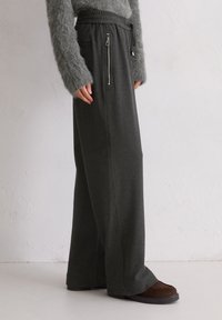Grijze wijde broek met een gladde textuur, voorzien van een ritszak en eenkoordsluiting, gecombineerd met donkere, platte schoenen.