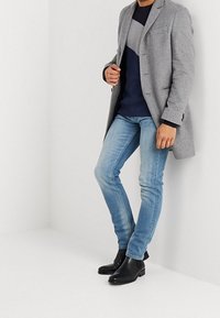 Cappotto di lana grigio su un maglione a strisce blu navy e grigio, jeans slim-fit azzurri e stivaletti neri, esposto su uno sfondo bianco.