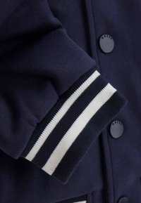 Giacca blu navy con una texture liscia, caratterizzata da polsini a costine a strisce bianche e blu navy e un davanti tinta unita. Include una semplice chiusura con bottoni.