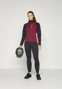 Vrouw in een kastanjebruine en zwarte fietsjack en zwarte legging die een grijze fietshelm vasthoudt, staande tegen een effen lichte achtergrond.