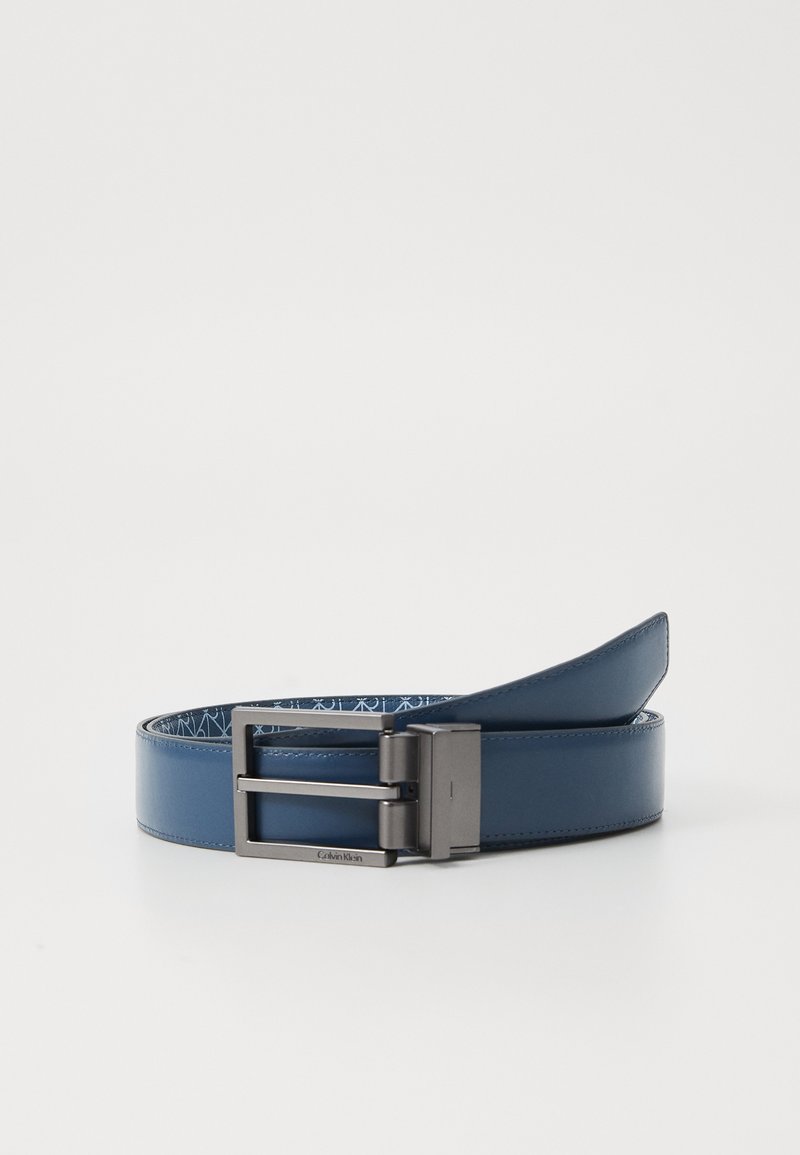 Ceinture en cuir bleu foncé avec boucle rectangulaire en métal de couleur gunmetal, partiellement enroulée, montrant un doublure intérieure à motif sur fond blanc uni.