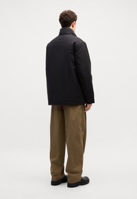 Giacca imbottita nera con colletto alto, vista da dietro. Abbinata a pantaloni beige chiaro, morbidi e larghi, e scarpe nere. Design semplice.