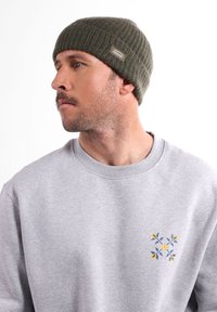 Bonnet en tricot vert olive à texture côtelée avec un patch logo, porté avec un sweat-shirt gris clair orné d'un motif floral bleu et jaune.