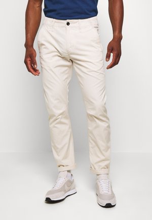 Homme portant un pantalon chino beige avec les revers roulés, des baskets blanches et grises, et une chemise bleu marine, debout devant un fond clair uni.