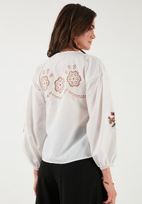 White blouse with puff sleeves, featuring intricate cut-out lace detailing and floral embroidery on the left sleeve. Smooth, lightweight fabric.