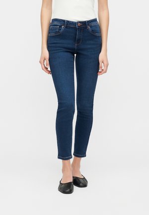 Femme portant un jean skinny bleu foncé et des ballerines noires à enfiler, debout devant un fond blanc uni.