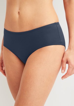 Vrouw draagt effen donkerblauwe bikinibroekjes, getoond van taille tot halverwege het bovenbeen tegen een effen achtergrond.