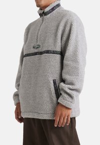 Grå fleecepullover med kvart lynlås, præget af mønstrede detaljer på kraven og brystet. Blød tekstur og afslappet pasform med sidelommer.