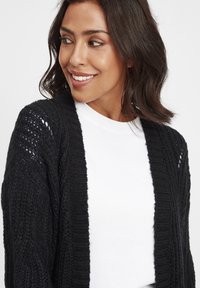 Svart virkad cardigan med öppen front, ribbat mönster och spetsliknande detaljer på ärmarna, bärs över en vit t-shirt.